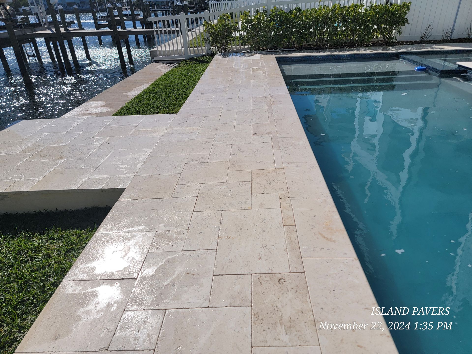 Stone pool deck beside a clear blue swimming pool, featuring waterfront views, lush grass, and marina railings.
