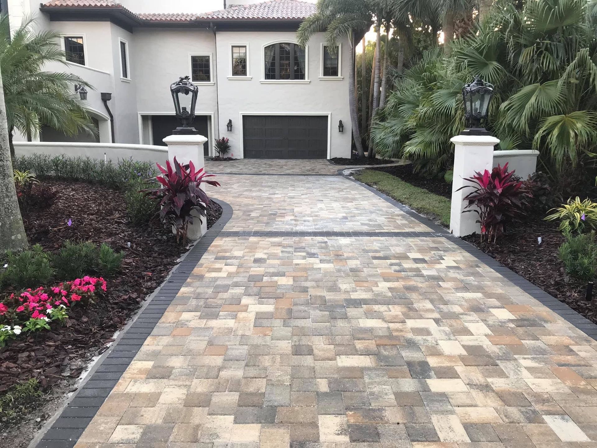 Paver driveway leading to a white house with a two-car garage and tropical landscaping