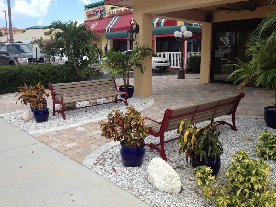 Outdoor shopping plaza with wooden benches, potted plants, and storefronts under an awning