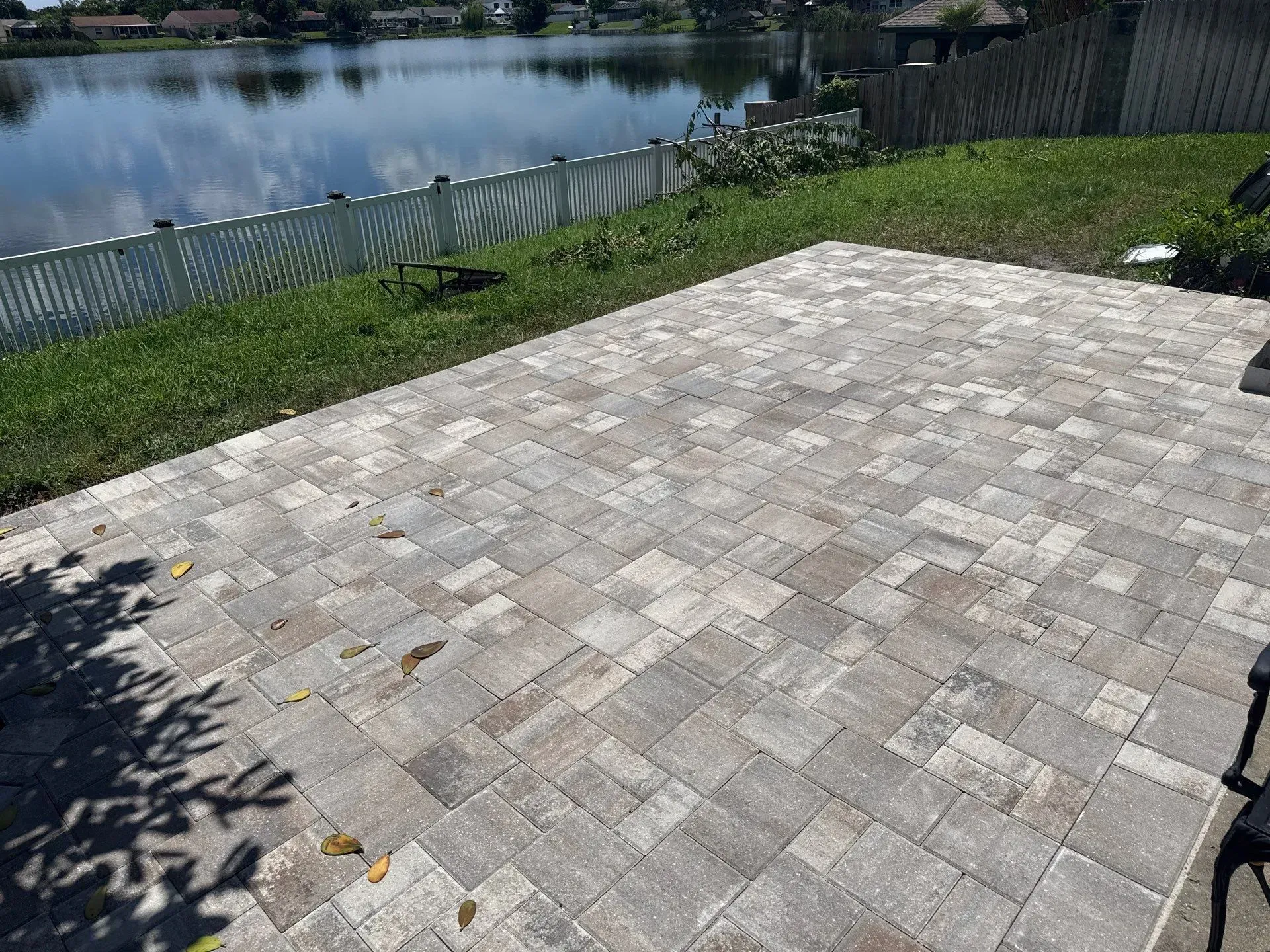 Wide brick paved driveway leading to a single-story gray house with green lawn and palm trees