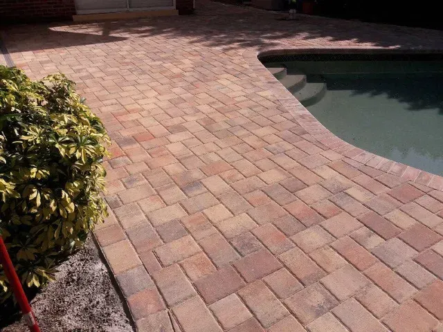 Brick paver patio beside a curved swimming pool, with a small shrub on the left.