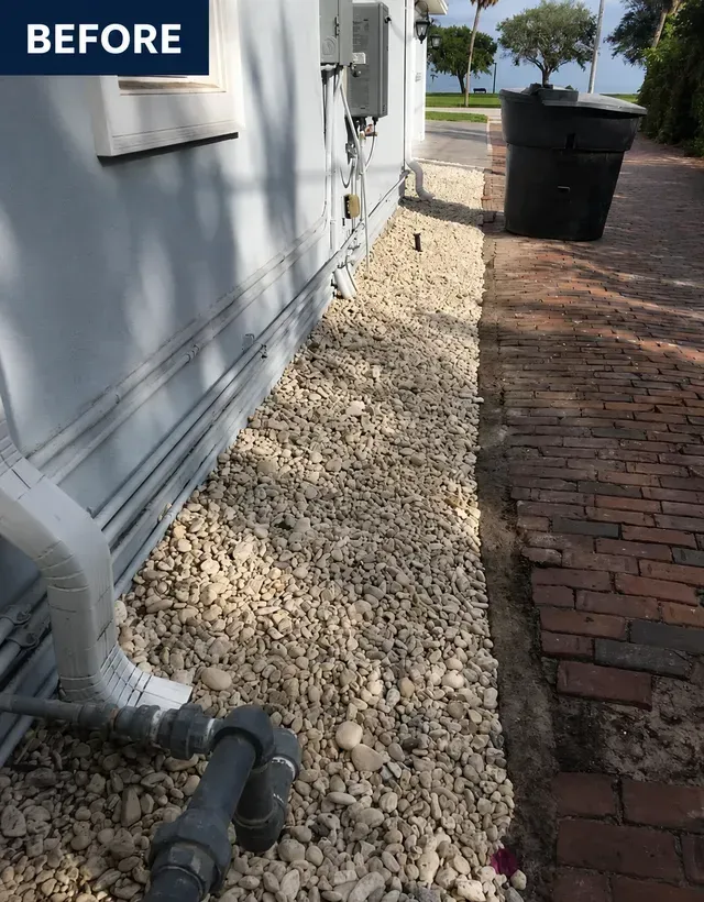 Side yard with white house wall, gravel bed, brick walkway, and trash bins in the background.