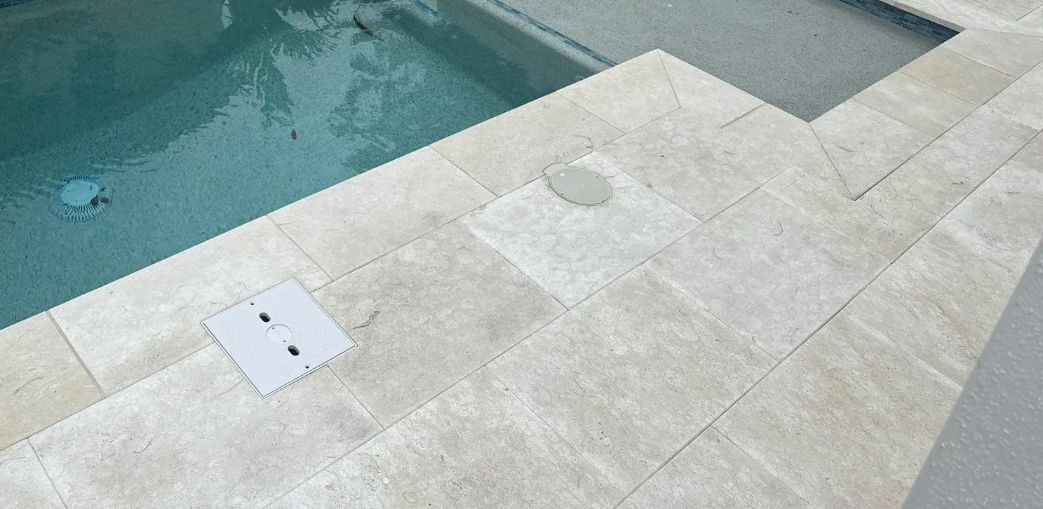 Travertine ivory stone pool deck beside turquoise water, viewed at a diagonal angle.