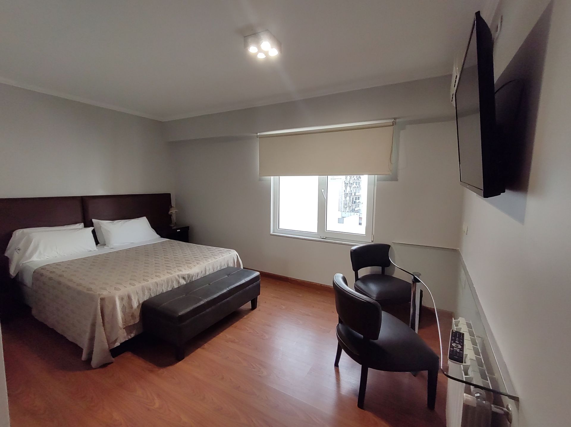 Bedroom with a bed, seating area, TV, and window. Wooden floors, neutral walls, and warm lighting.