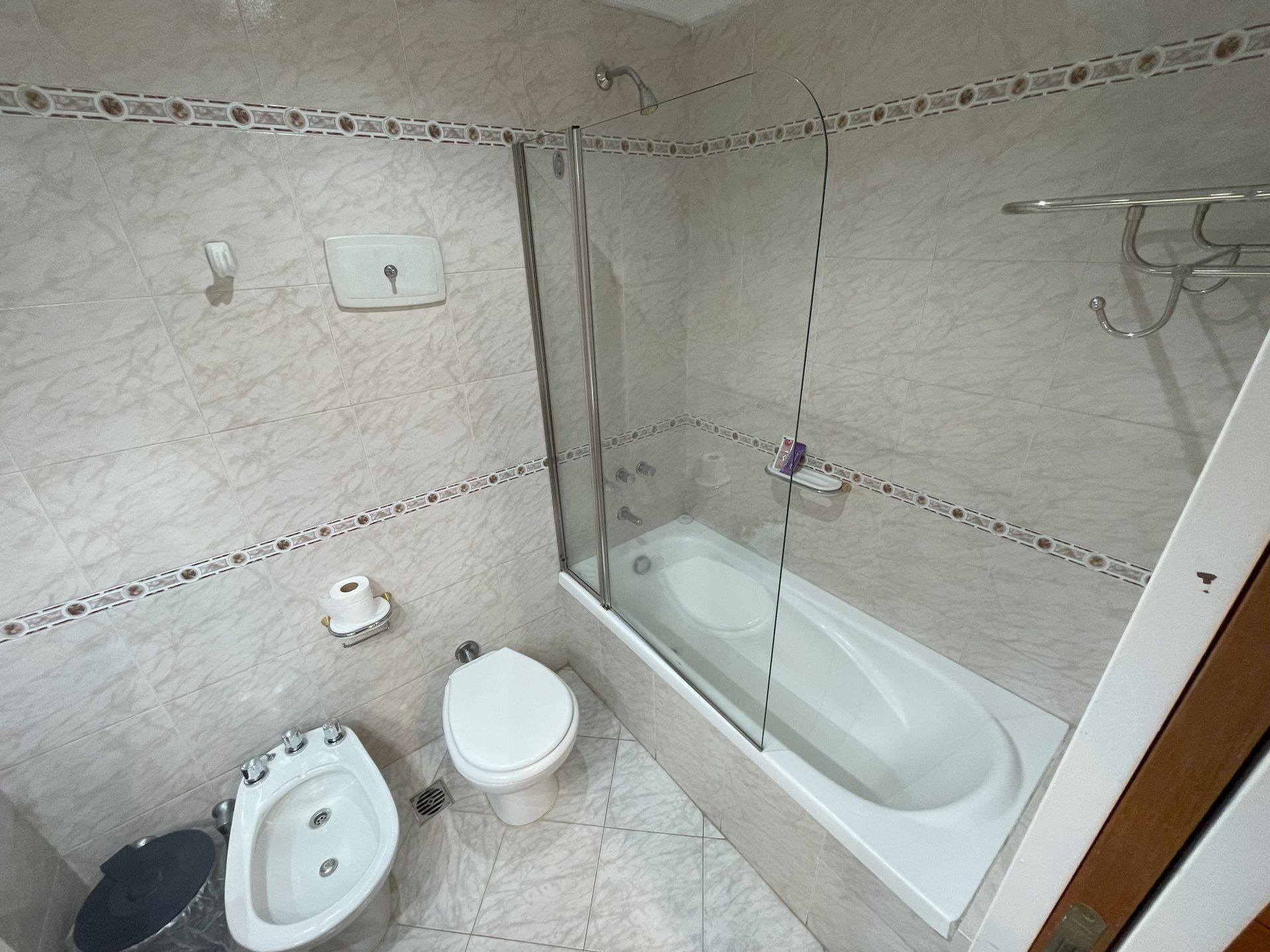 Baño pequeño de color beige con inodoro, bidé y bañera con paredes de cristal.