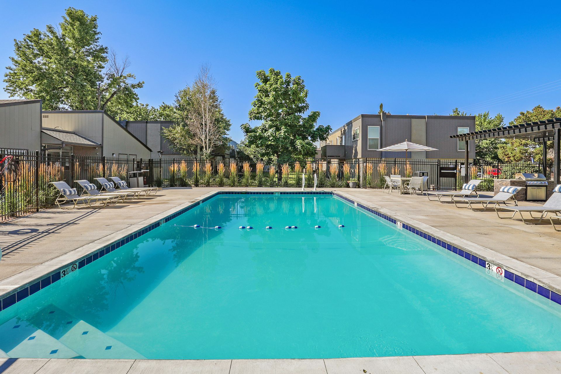 A sunny, rectangular swimming pool surrounded by lounge chairs, patio furniture, and apartment buildings.