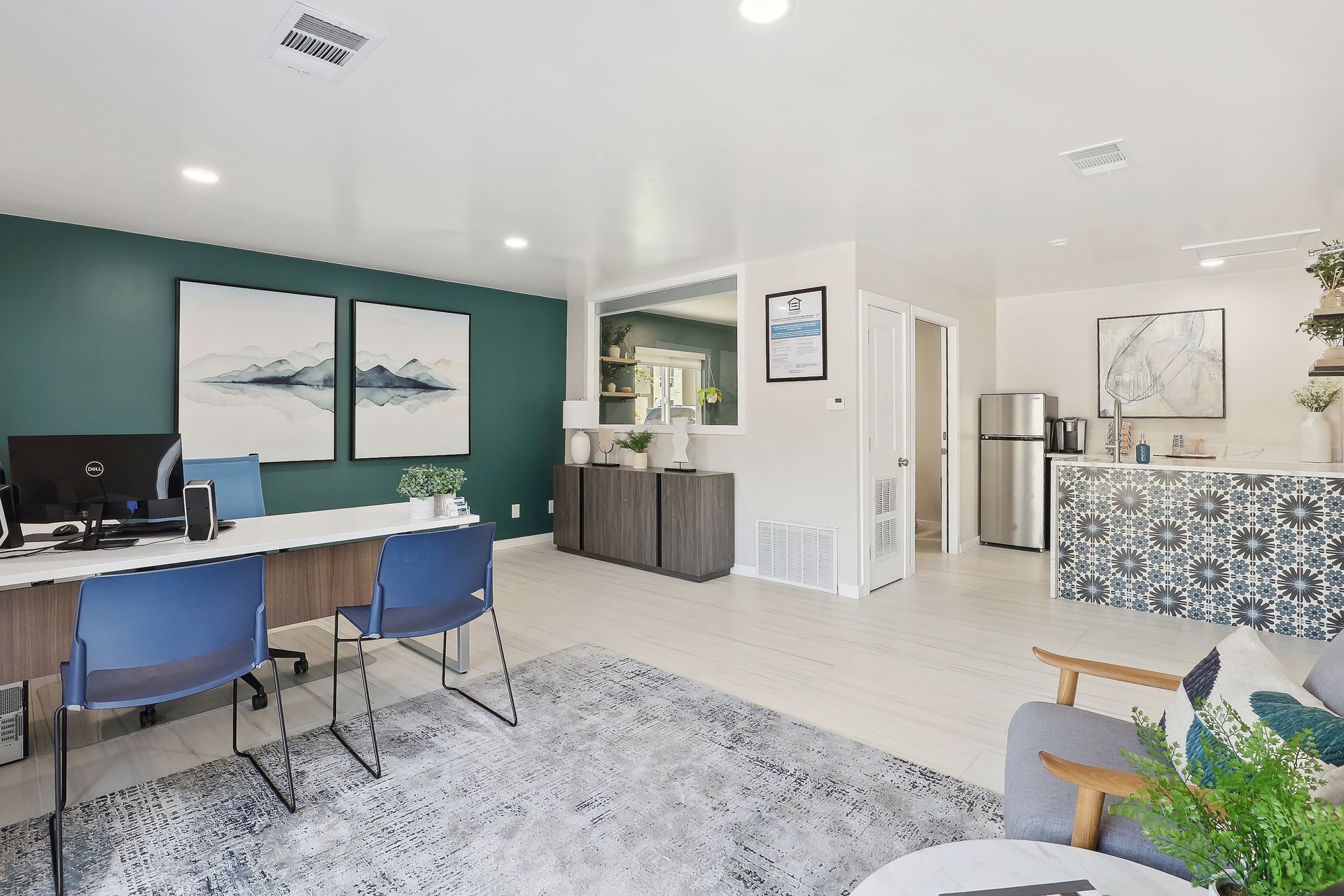 A bright office space with a teal accent wall, two desks with blue chairs, a kitchen area, and a patterned counter.