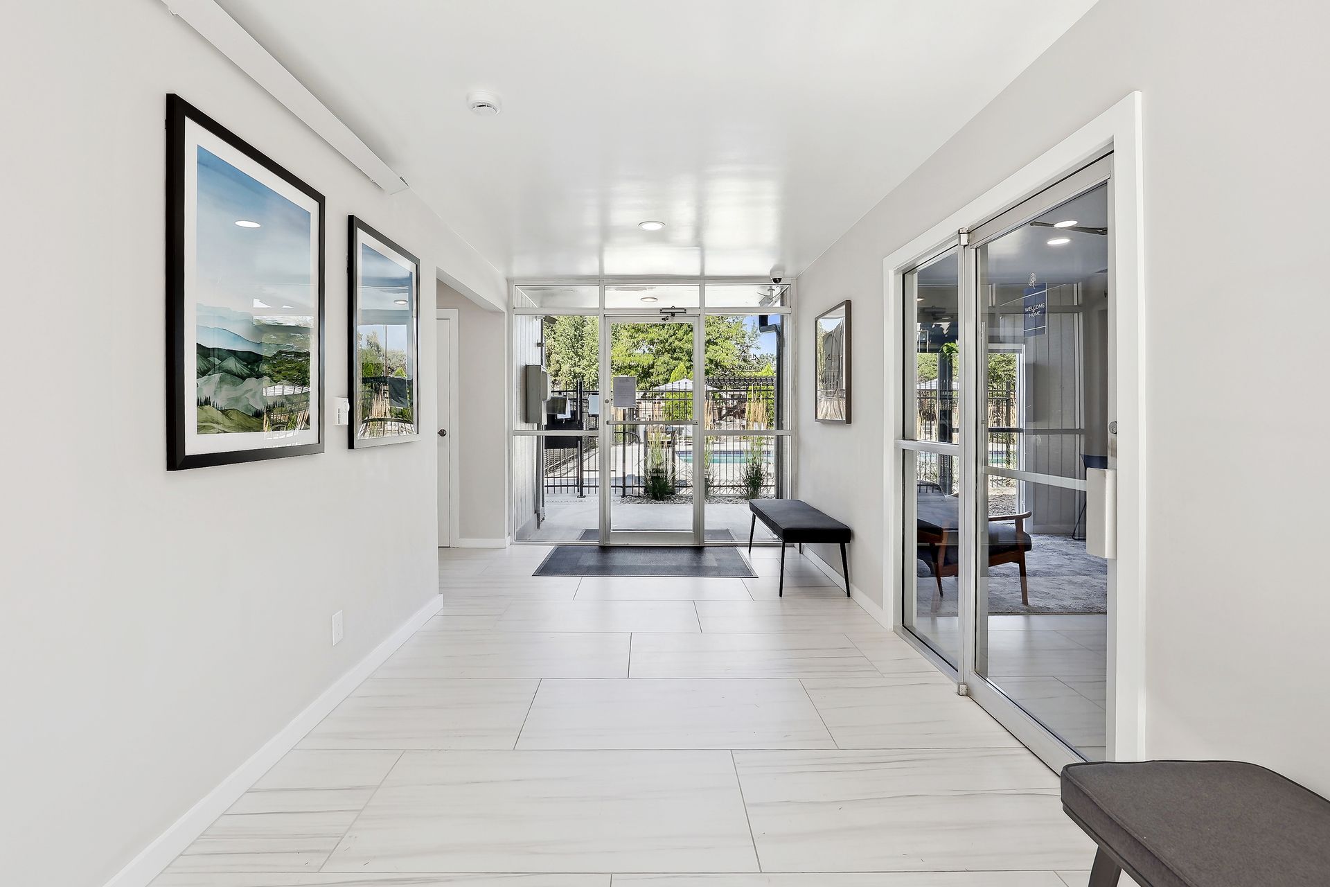 A brightly lit, minimalist hallway with white floors leads to glass doors, with framed art on the wall and benches.