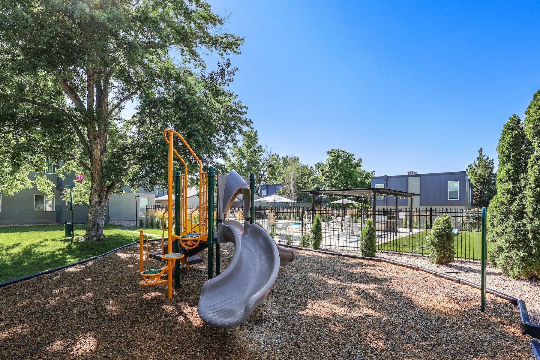 A playground with a yellow frame and curved gray slide on wood chips, with a pool and apartment building in the distance.
