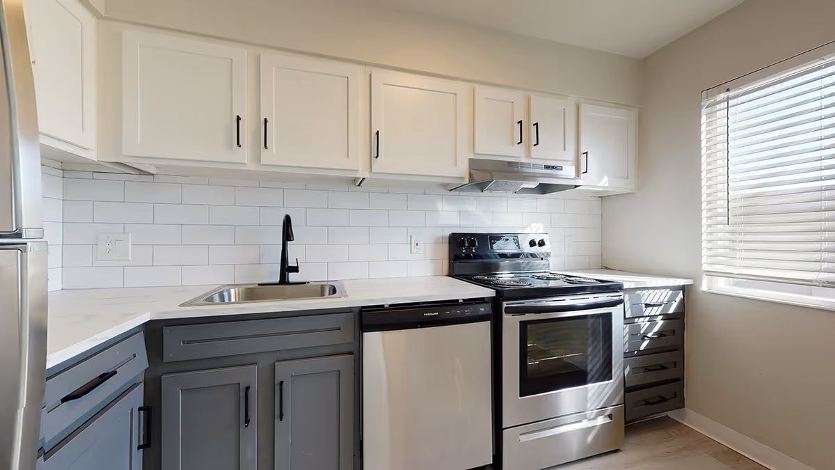 A kitchen with white upper cabinets, gray lower cabinets, white tile backsplash, stainless appliances, and a window.