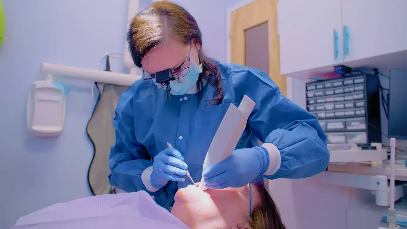 Clinician in magnifying loupes examining a reclined patient's teeth at a consultation before an extraction and socket graft