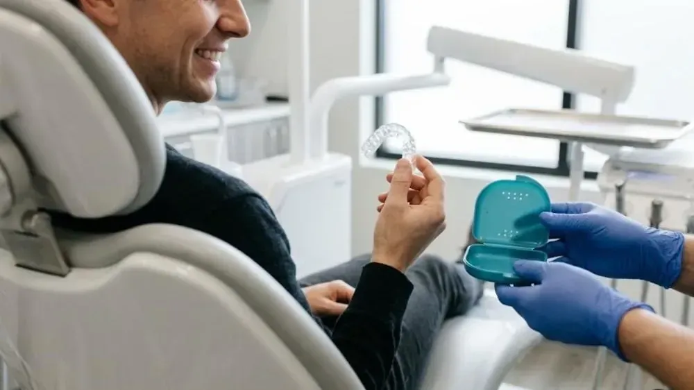 Smiling patient in the dental chair holding his new clear custom nightguard as gloved hands offer its case