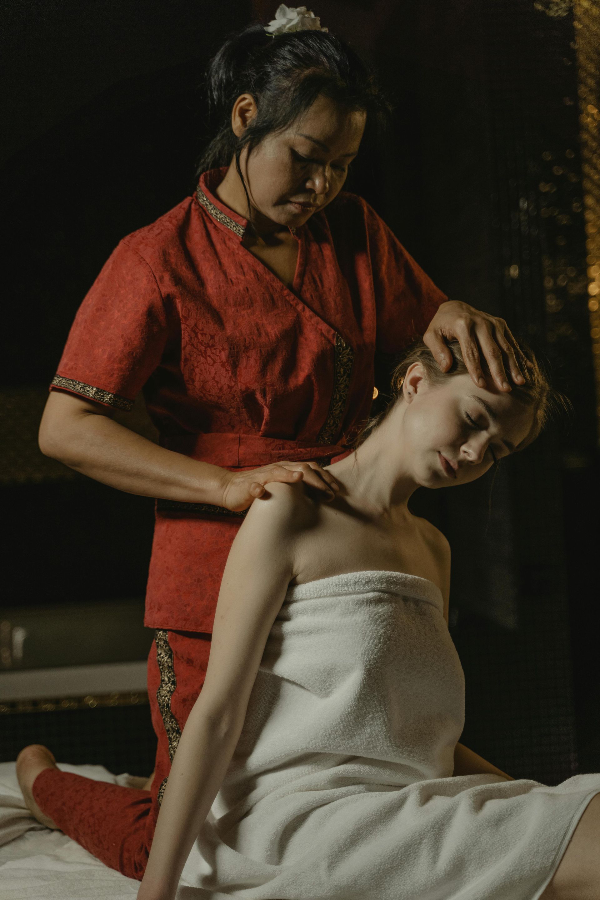 A massage therapist in red scrubs performs a neck and shoulder stretch on a person wrapped in a white towel.