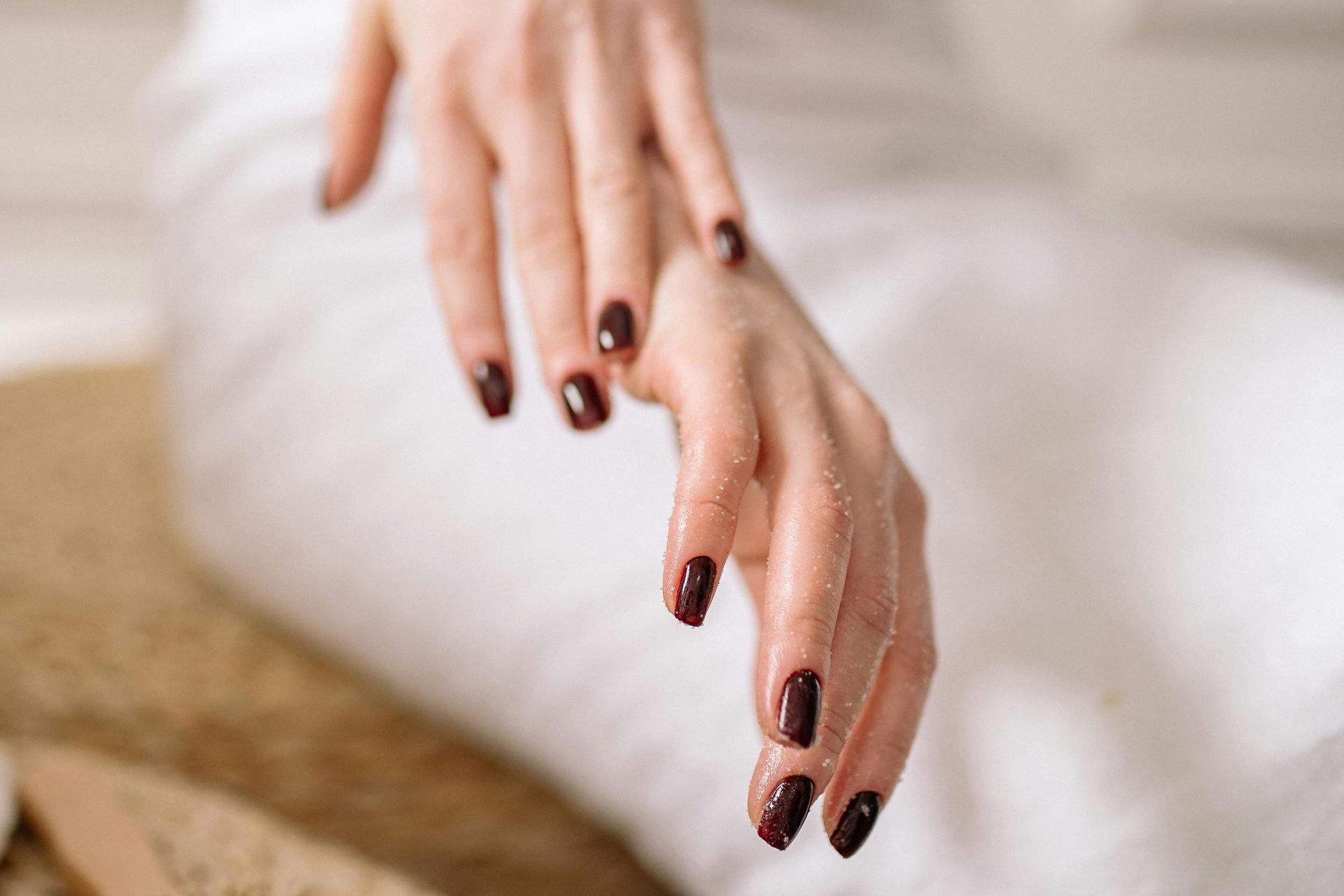 Hands with dark manicured nails rest gracefully against a textured, light-colored fabric.