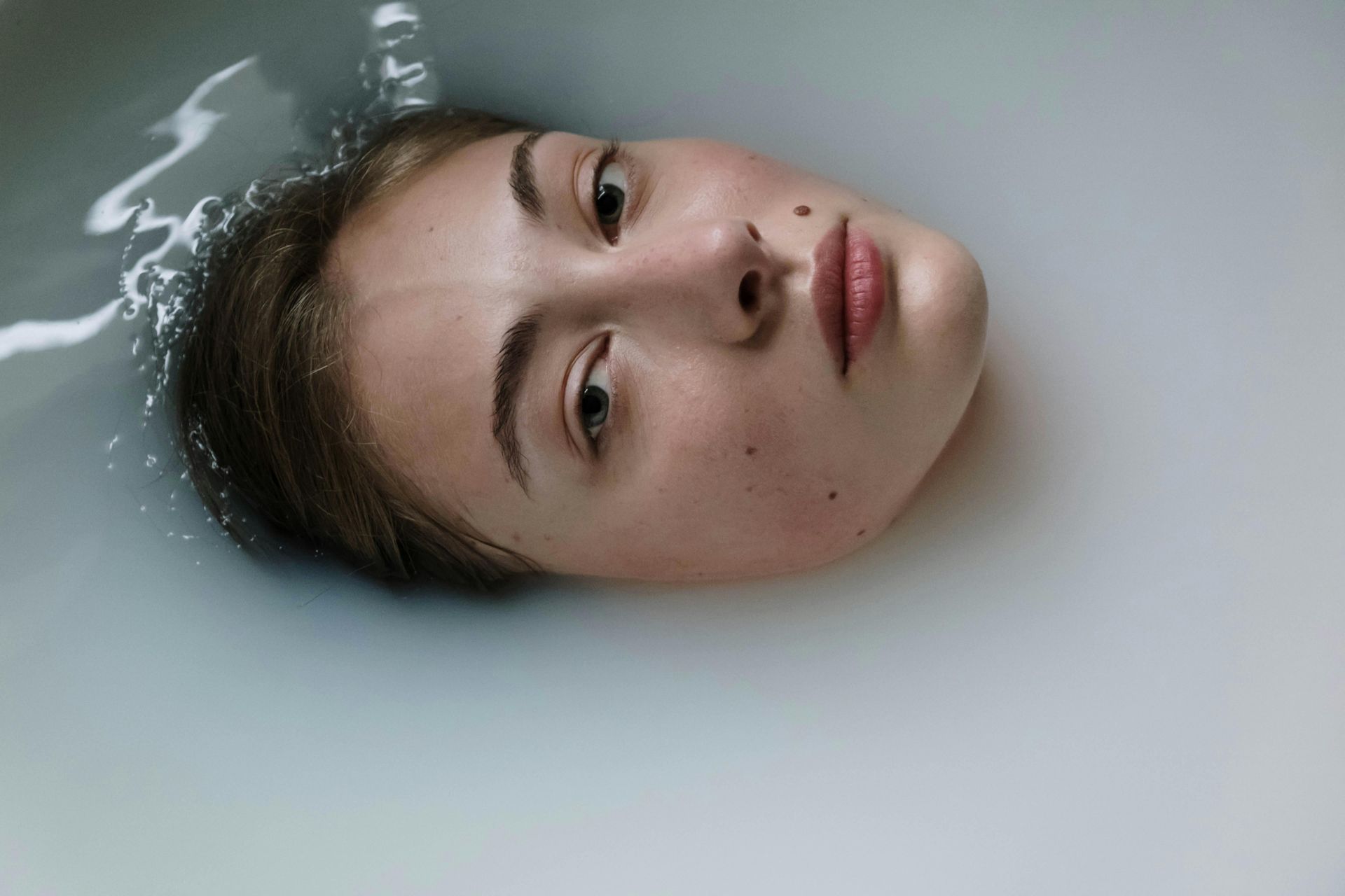A person’s head and face floating in milky, opaque liquid, looking upward with a neutral expression.