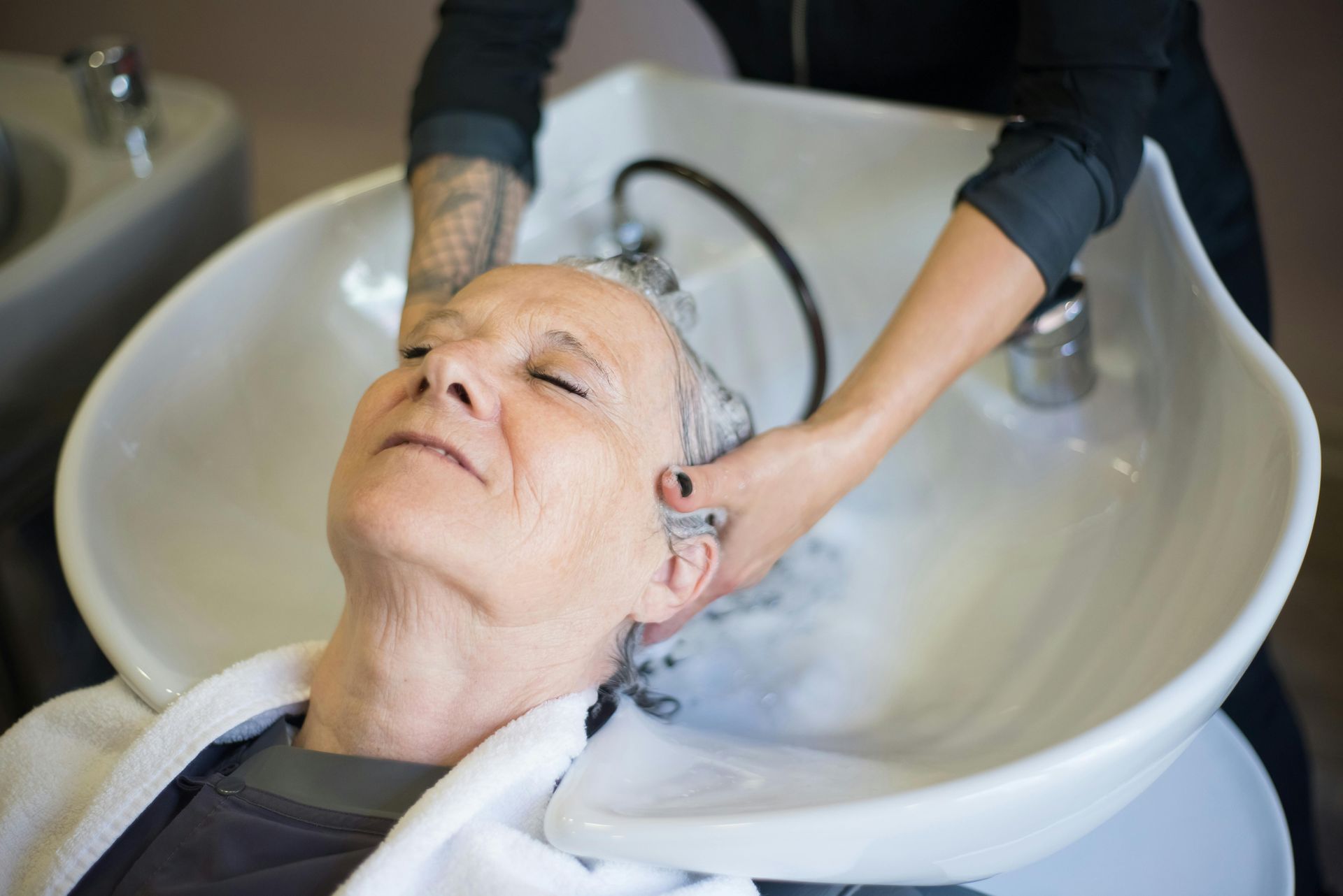 A professional stylist washes the hair of a relaxed person reclined in a salon sink.