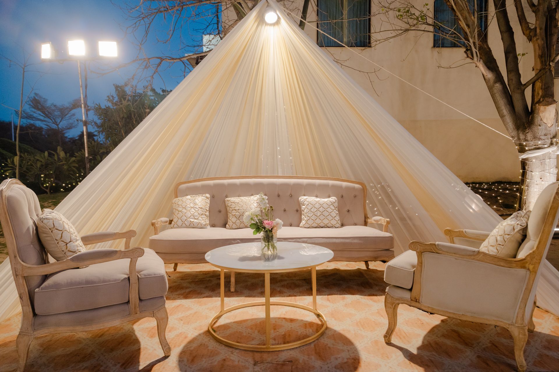 Outdoor patio lounge with canopy drapes, cushioned sofas, round table, and warm string lights at dusk
