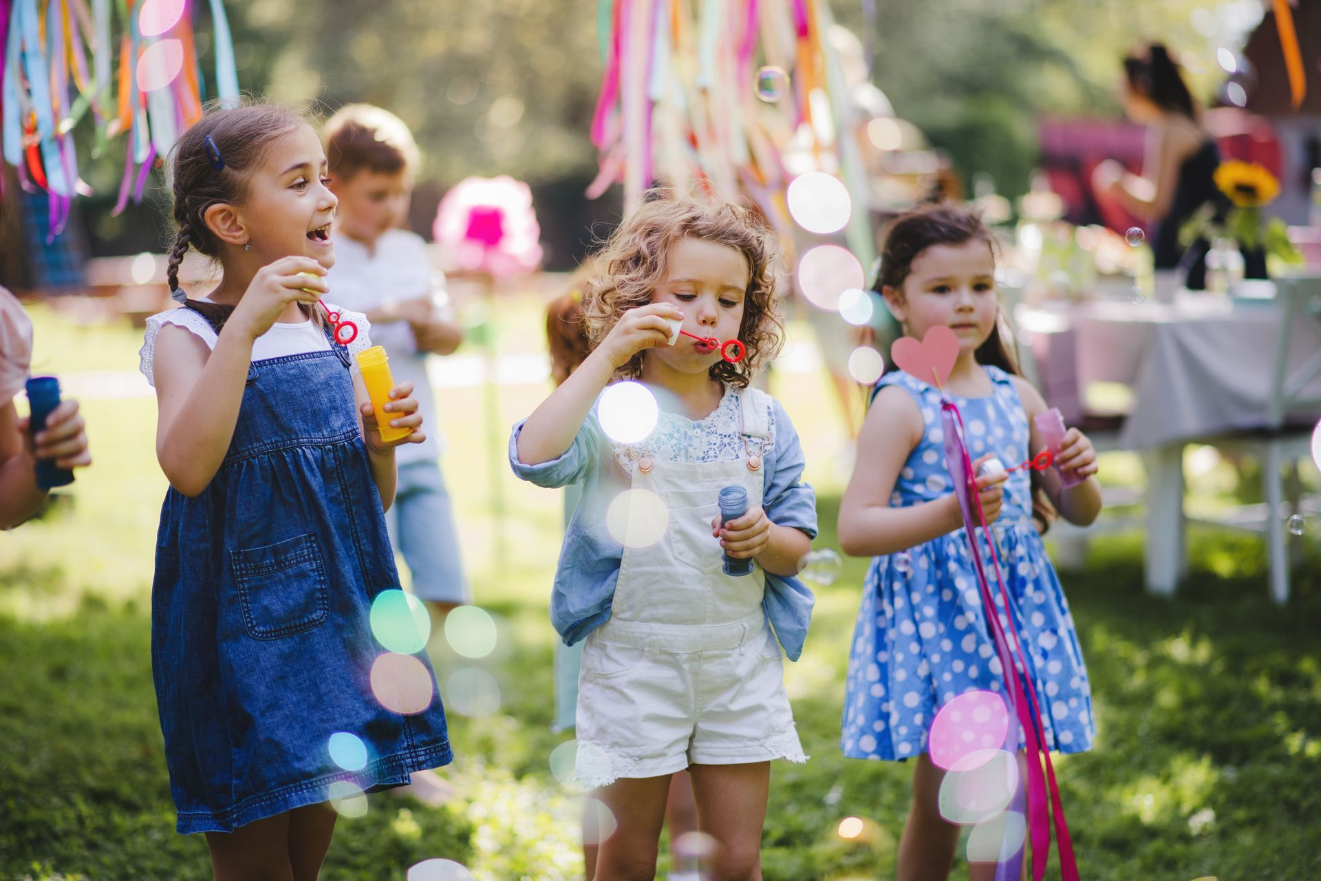 Children blowing bubbles at an outdoor party, with colorful streamers and a festive table in the background