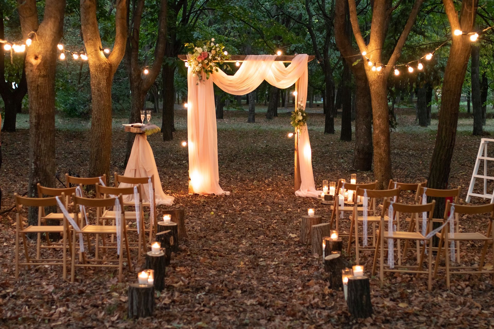Outdoor wedding aisle with white draped arch, string lights, and wooden chairs among trees at dusk