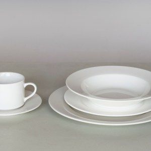 White cup and saucer beside stacked white plates and a bowl on a gray surface