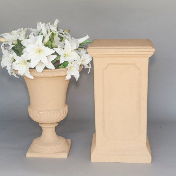 Beige decorative urn with white flowers beside a matching pedestal on a gray background