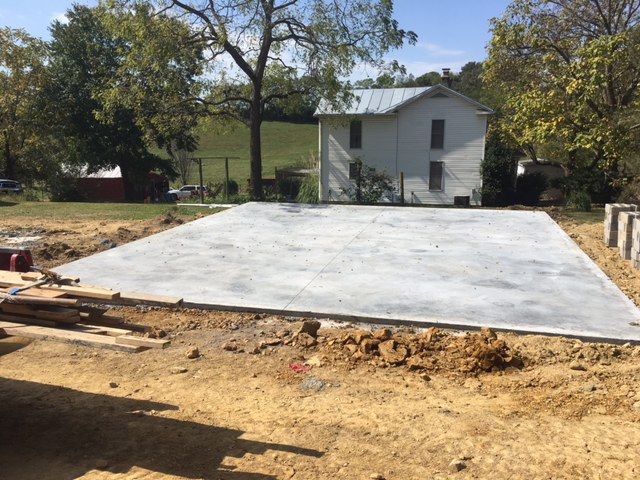 A freshly poured concrete slab foundation sits in a dirt-filled lot with a white house in the background.