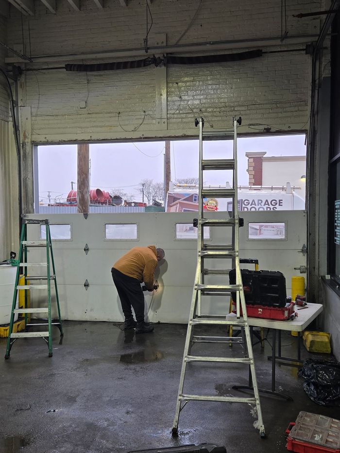 Man working on a garage door inside a building. Ladder and tools present. Snow visible outside.