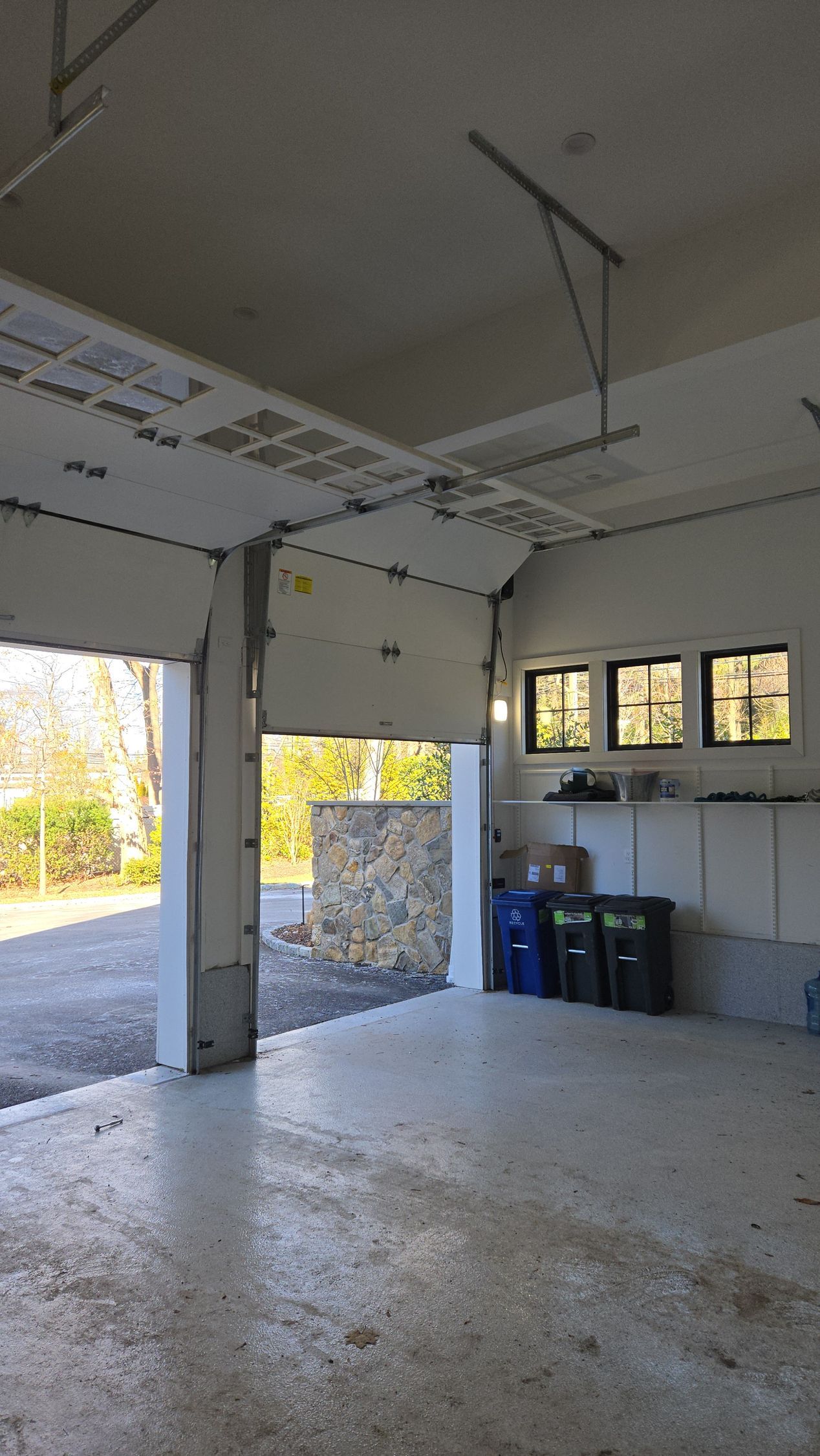 Inside a garage, the garage door is open, showing a driveway and foliage outside.