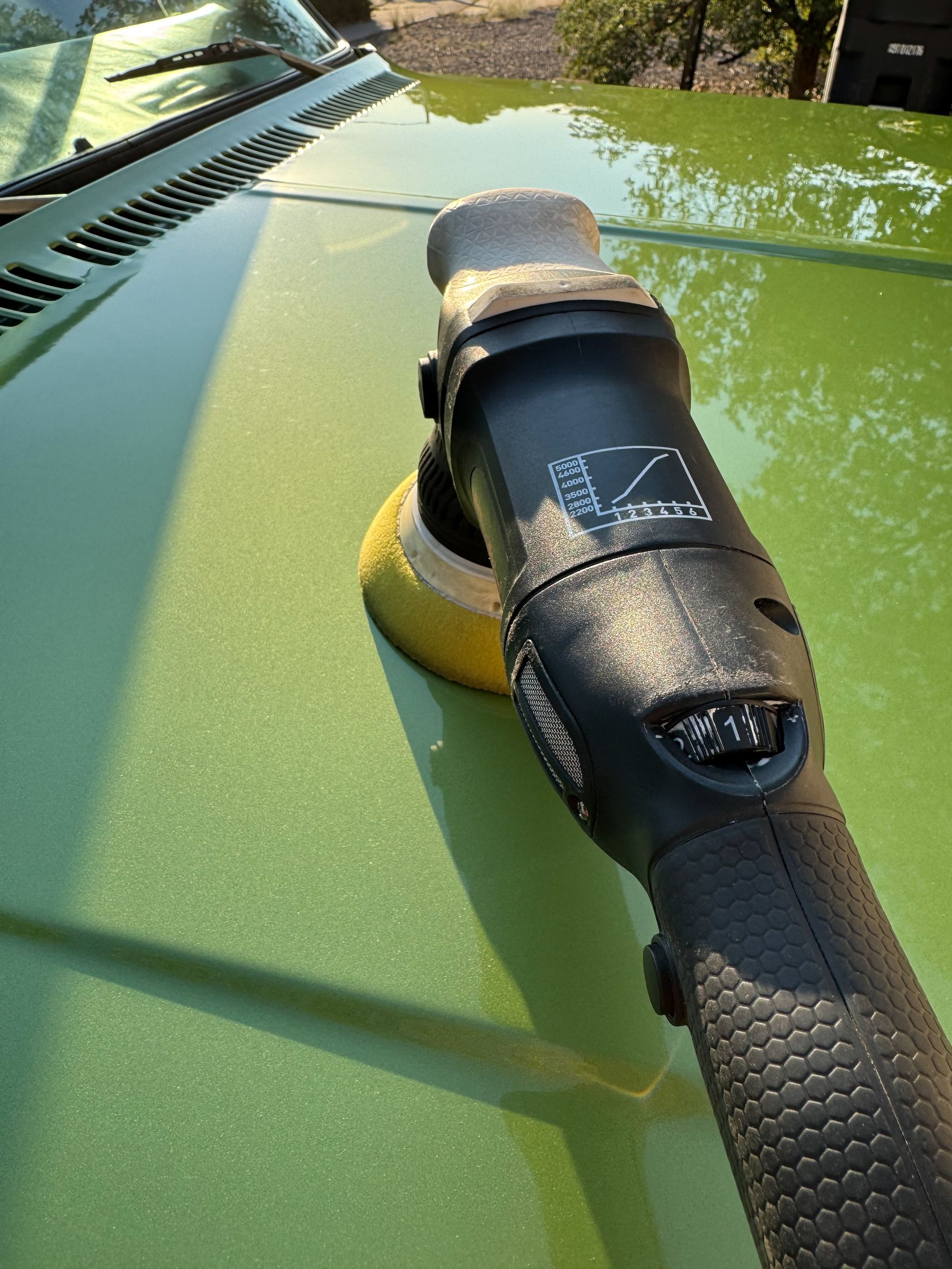 Polishing machine on a green car hood, reflecting sunlight.