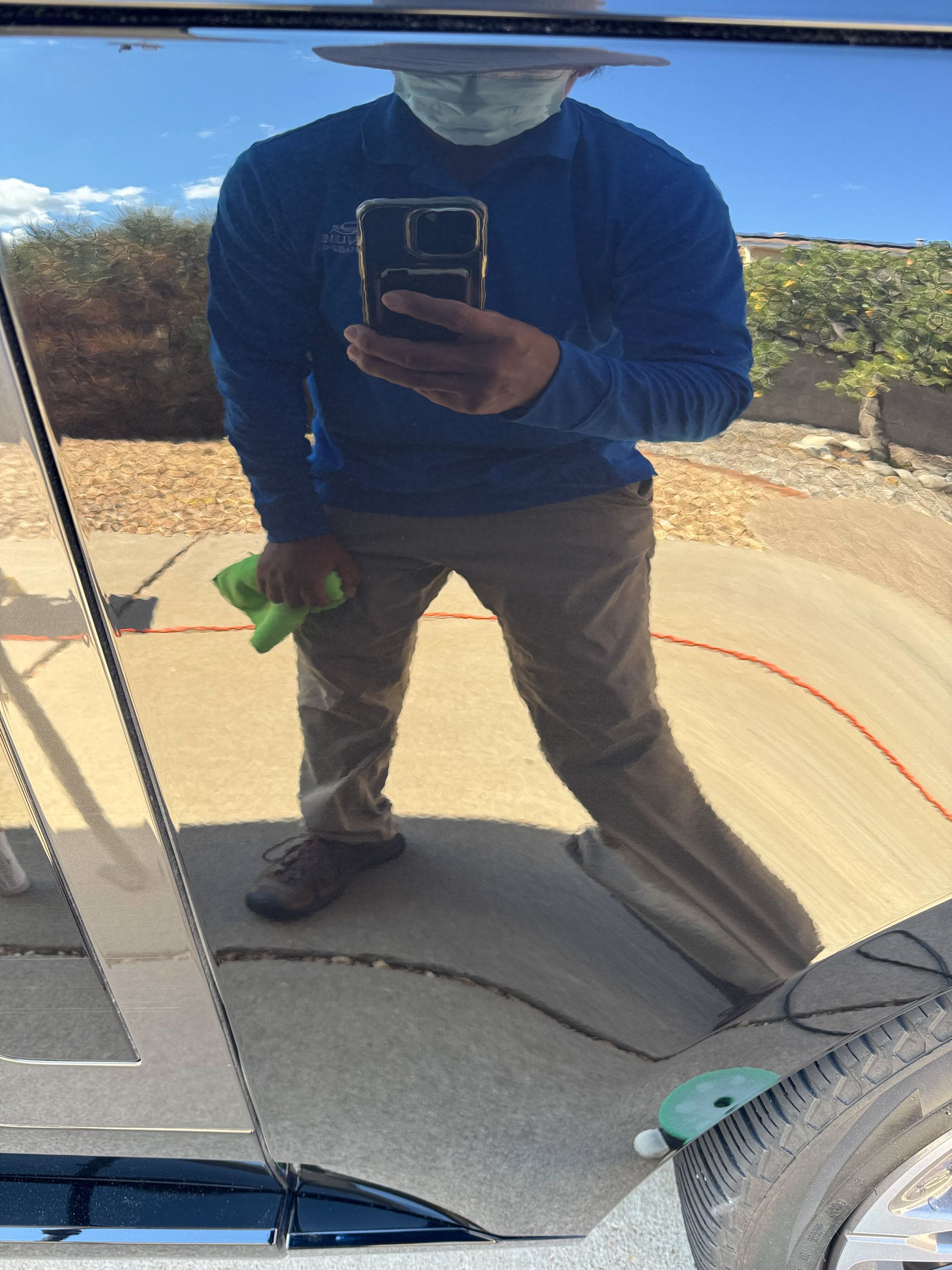 Person in blue shirt and mask reflecting in car, holding green cloth. Brown pants, blue sky.