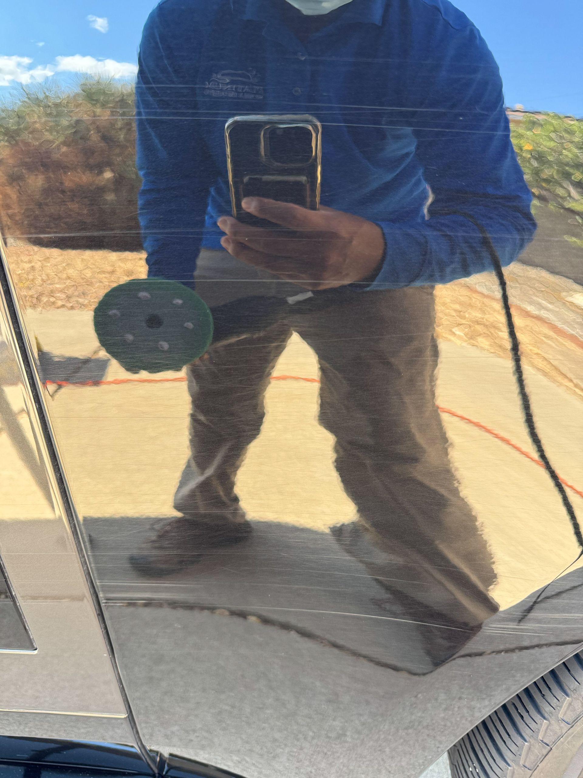 Man in blue shirt polishing a car, holding tool, reflection of sky and surroundings in car surface.
