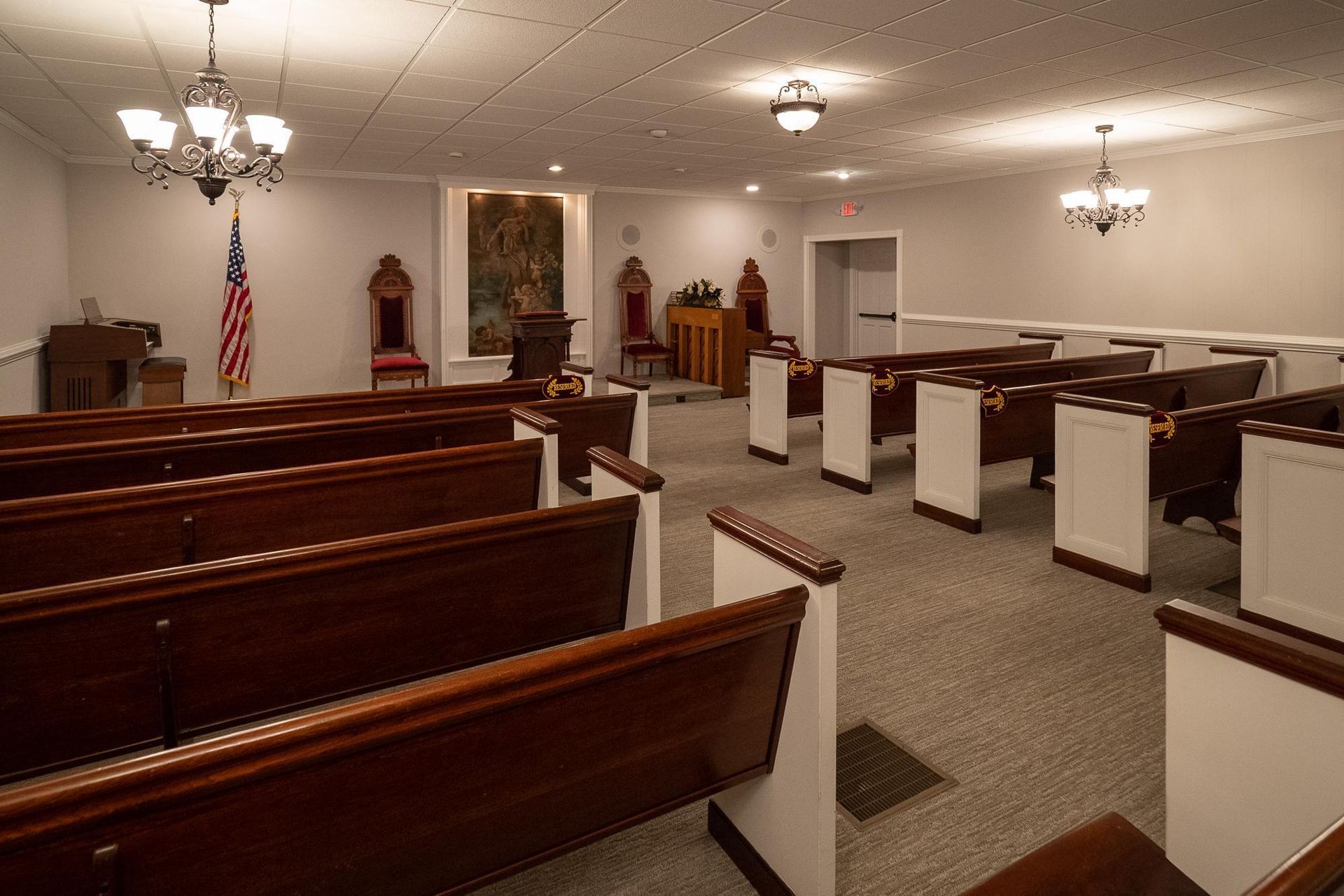 Empty church sanctuary with wooden pews, white railings, chandeliers, and an American flag near the front.