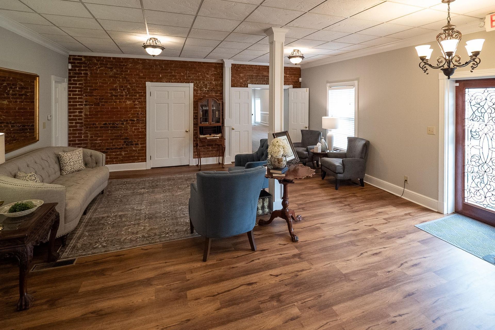 Spacious living room with wood floors, brick wall, blue chairs, sofa, and a glass door to outside.