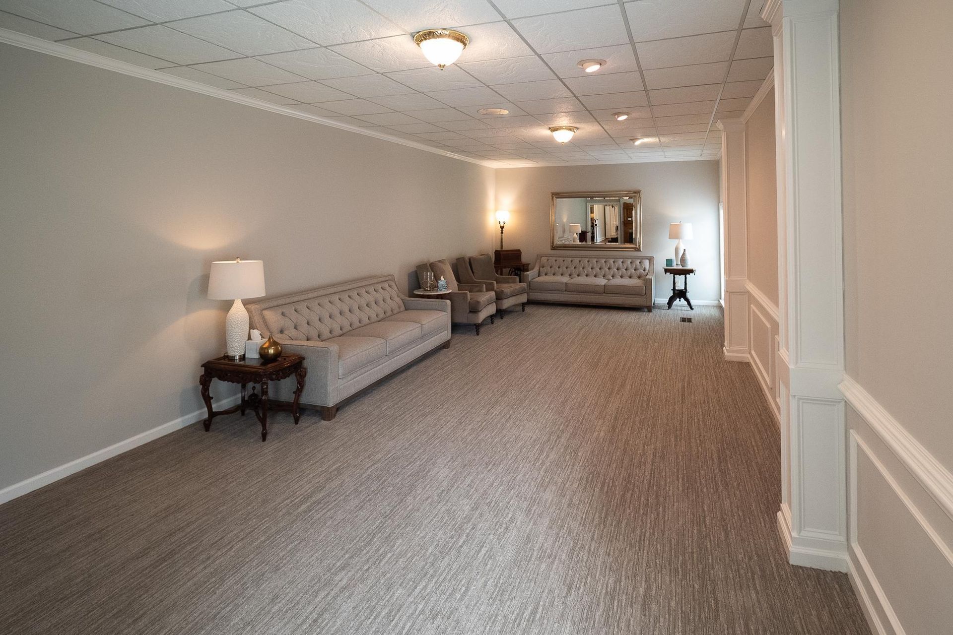Empty carpeted living room with gray sofas, side tables, and wall mirror under ceiling lights
