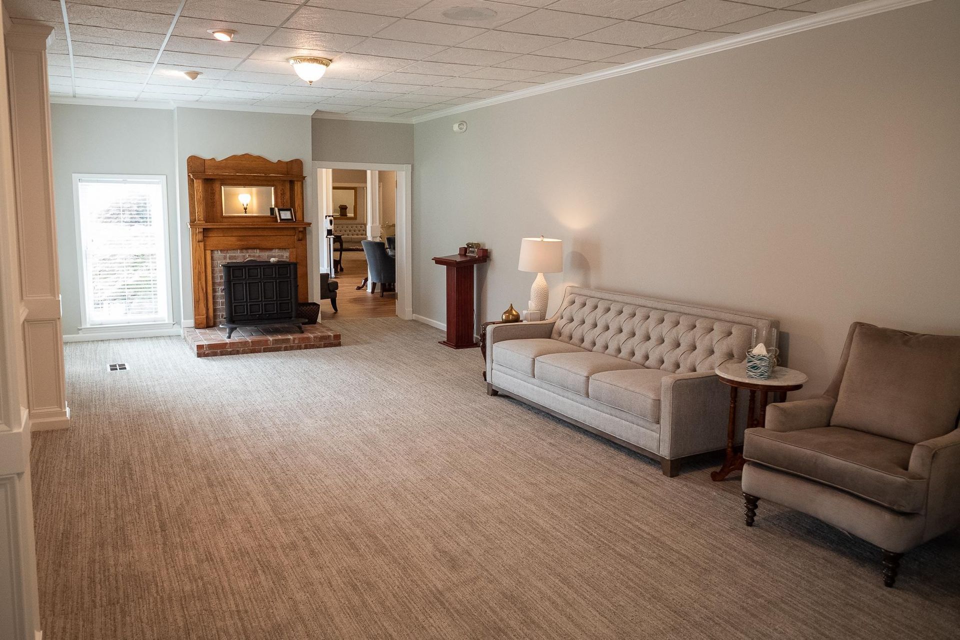 Spacious beige living room with tufted sofa, armchair, lamp, fireplace, and carpeted floor