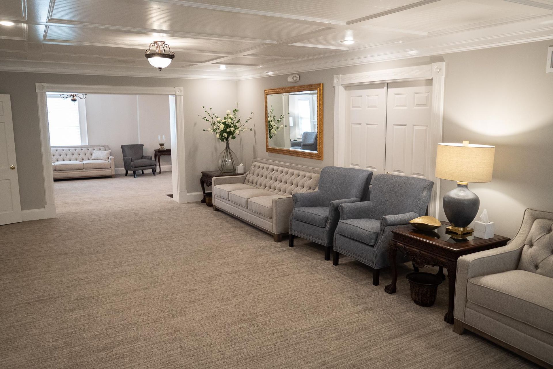 Spacious beige living room with sofa, armchair, side table, lamp, mirror, and open doorway to bedroom