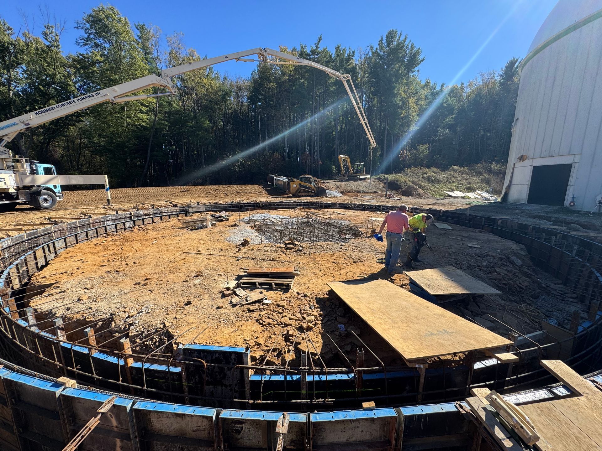 Construction site with circular foundation, concrete truck, and worker beside a white building under clear sky