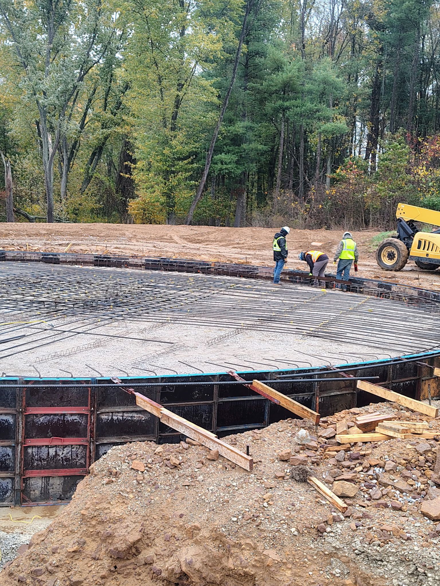 Construction workers and a yellow excavator on a concrete bridge foundation in a wooded area
