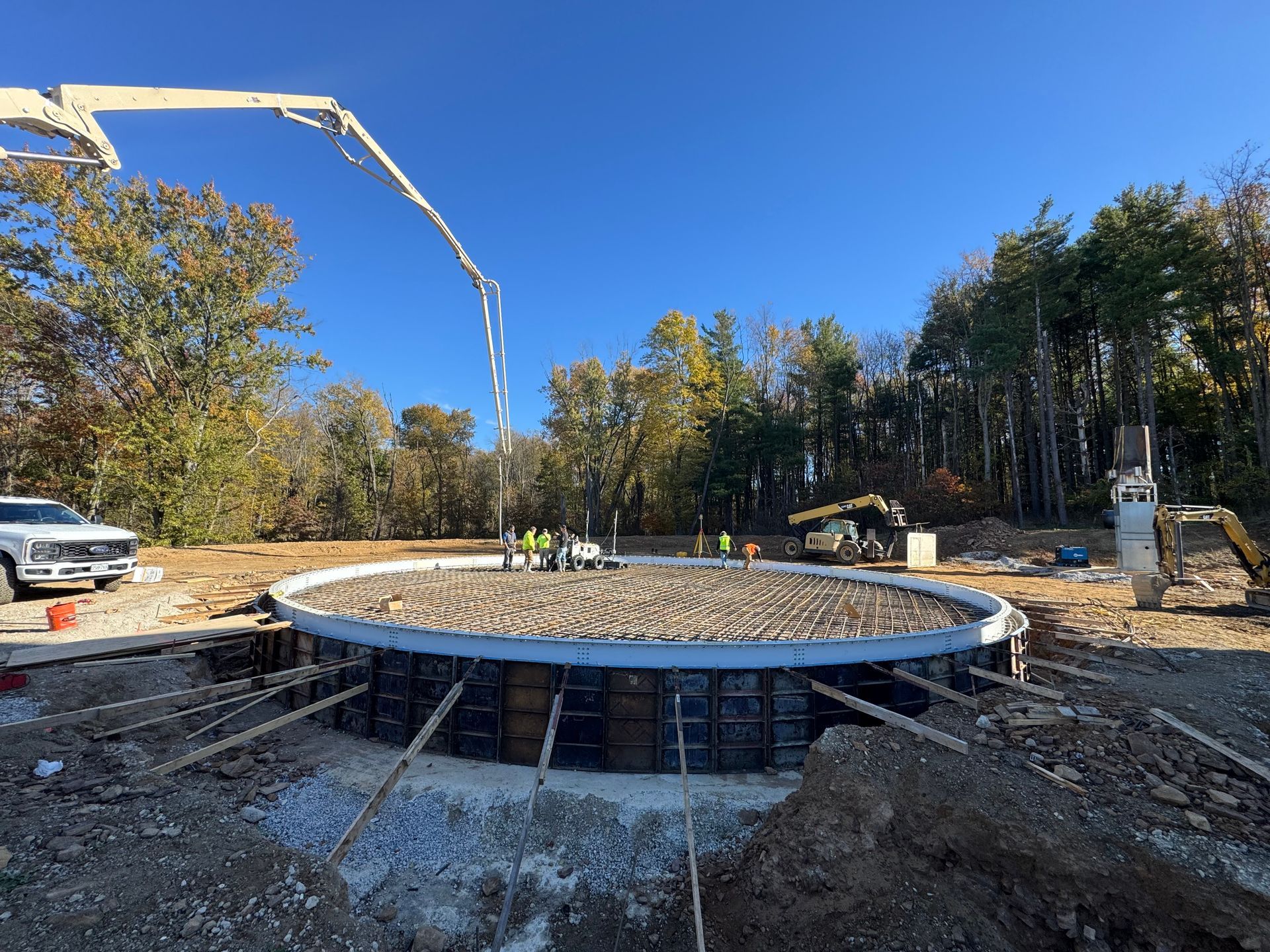 Circular concrete foundation under construction with workers, truck, and pump hose in a wooded lot