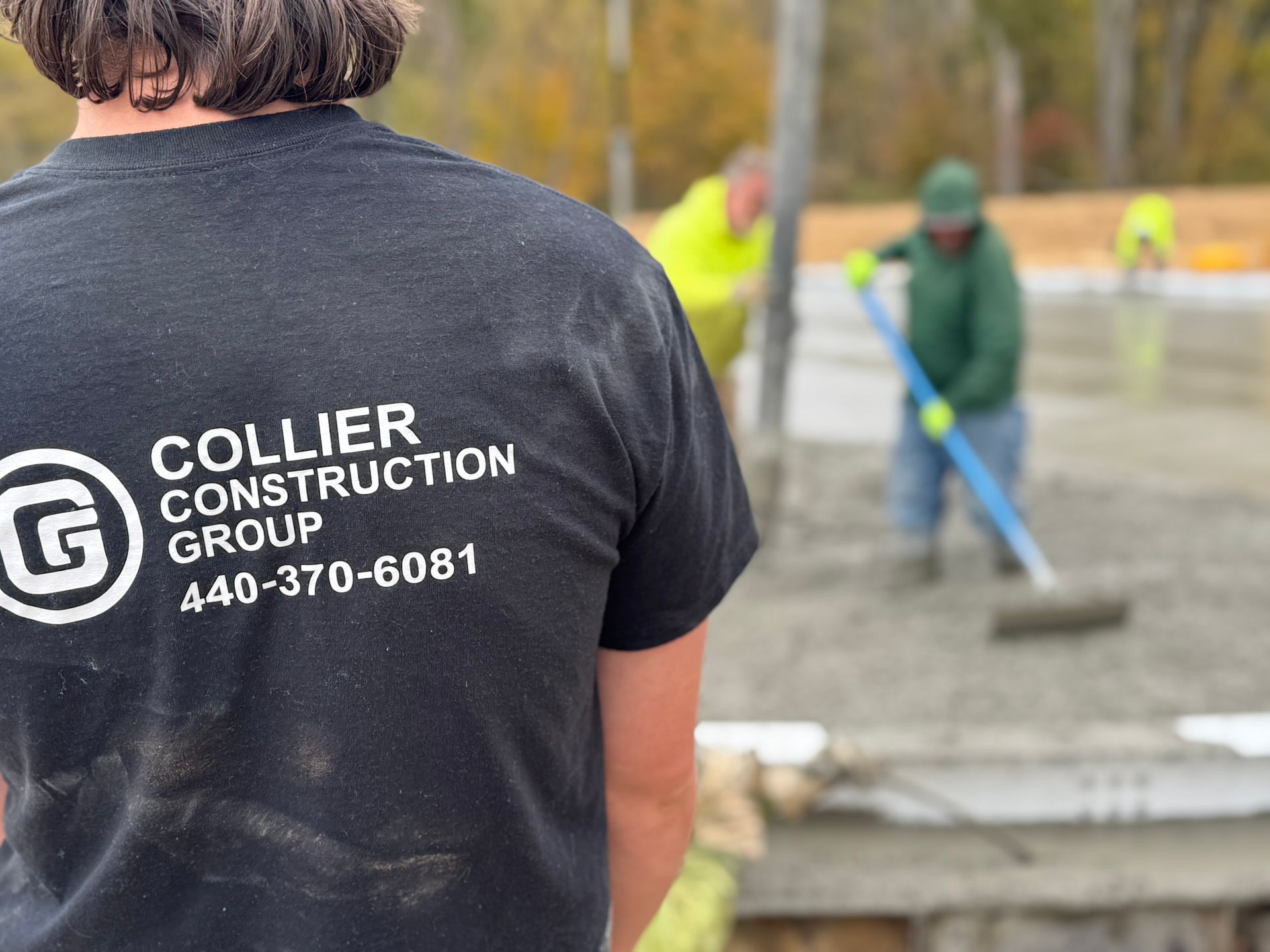 Worker in dark shirt at construction site, with crew smoothing wet concrete in the background.