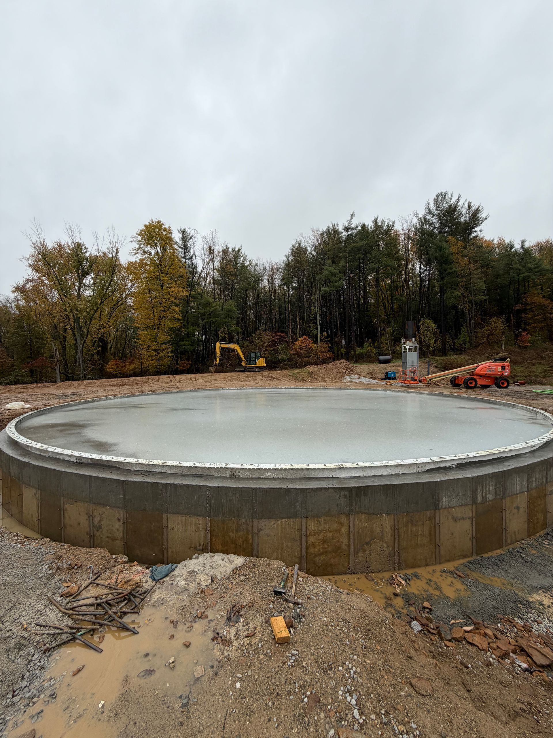 Large circular concrete tank under construction in a muddy outdoor site with excavator and trees in the background