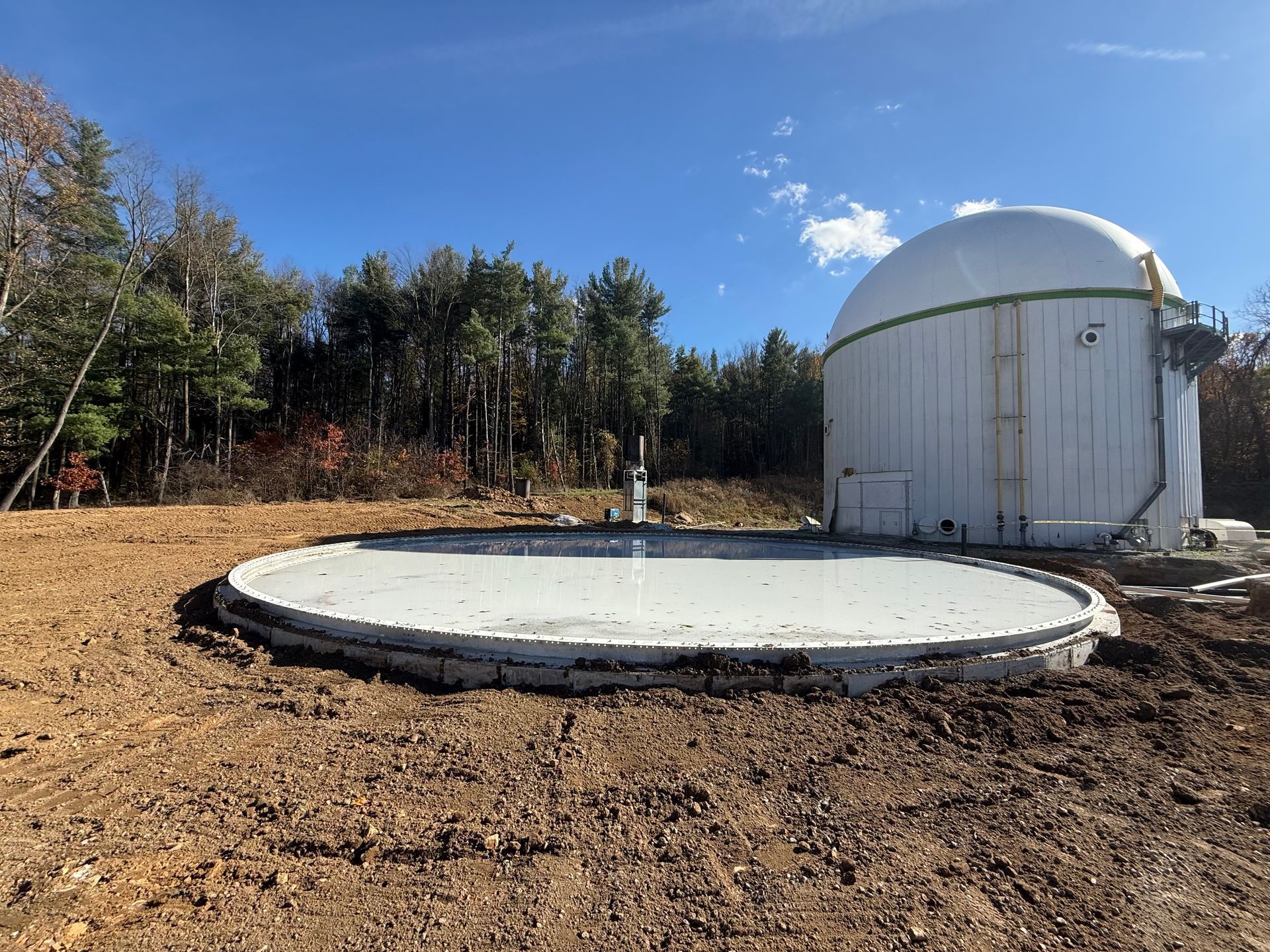 Circular concrete foundation beside a white dome-topped utility building in a cleared wooded area under blue sky