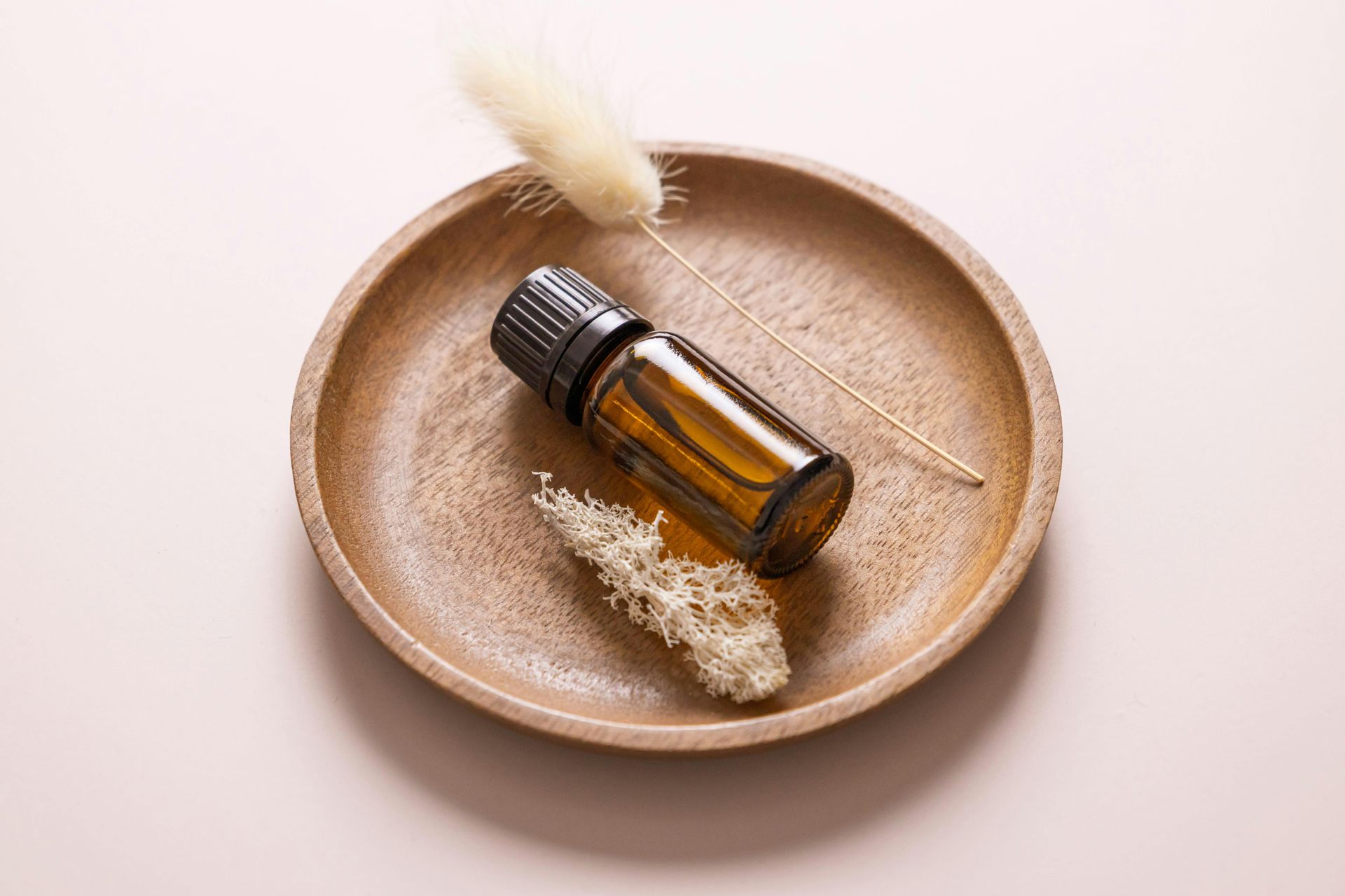 Amber essential oil bottle on a wooden plate with dried plants, set on a pink background.