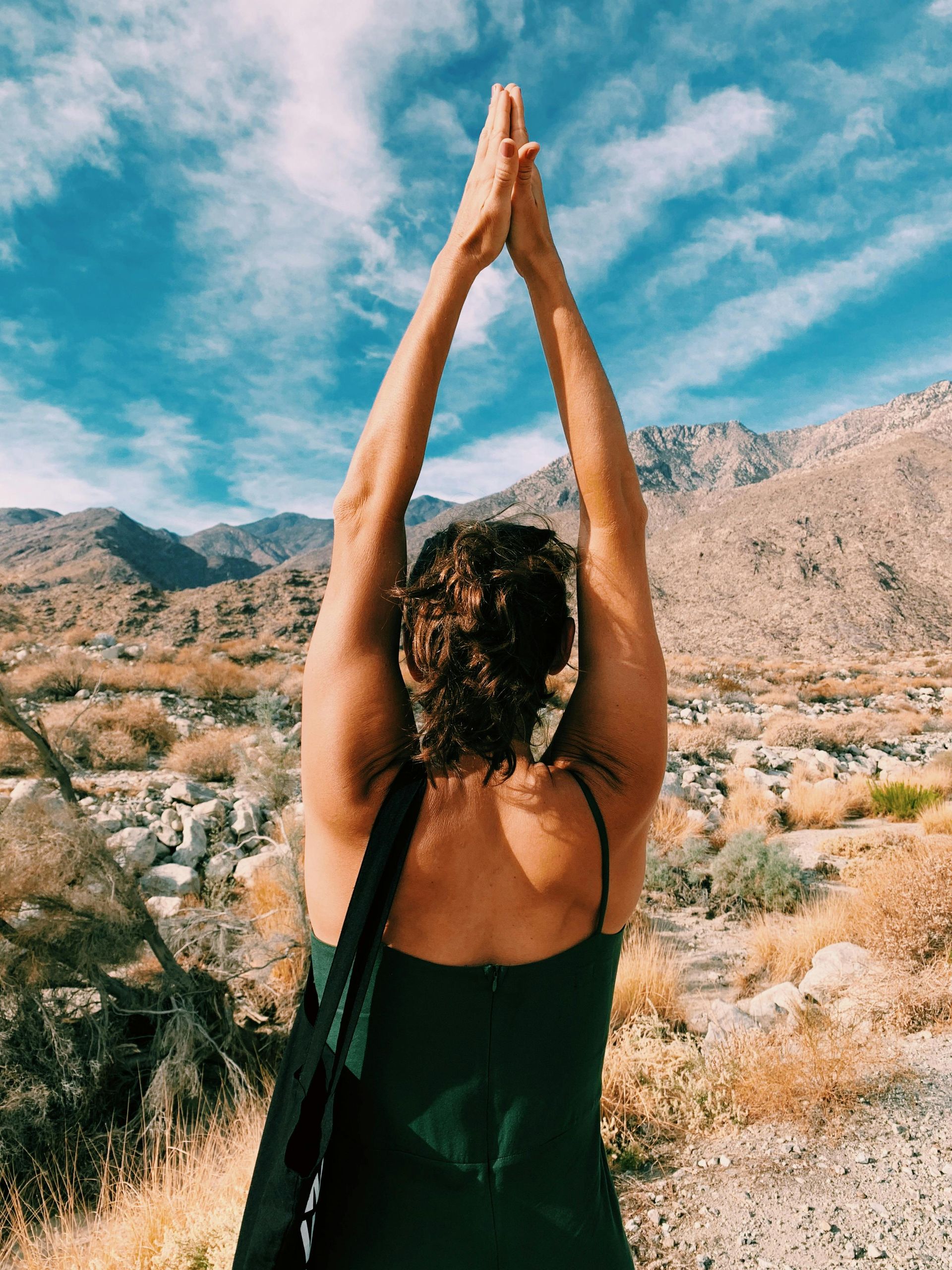 Woman with arms raised in prayer pose, facing mountains, wearing a dark green dress, sunny outdoor setting.