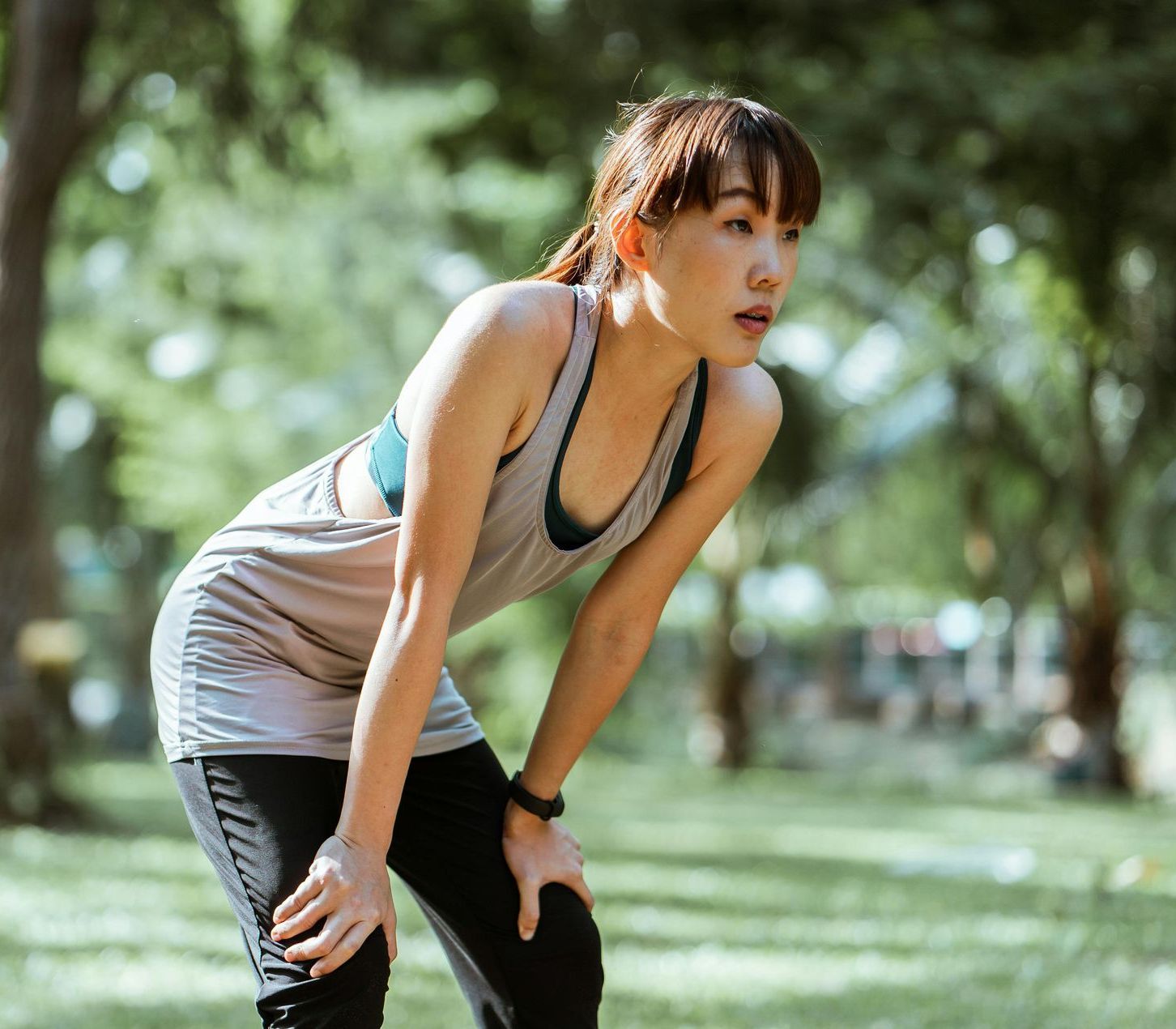 Woman in athletic wear, breathing heavily with hands on knees in a park.