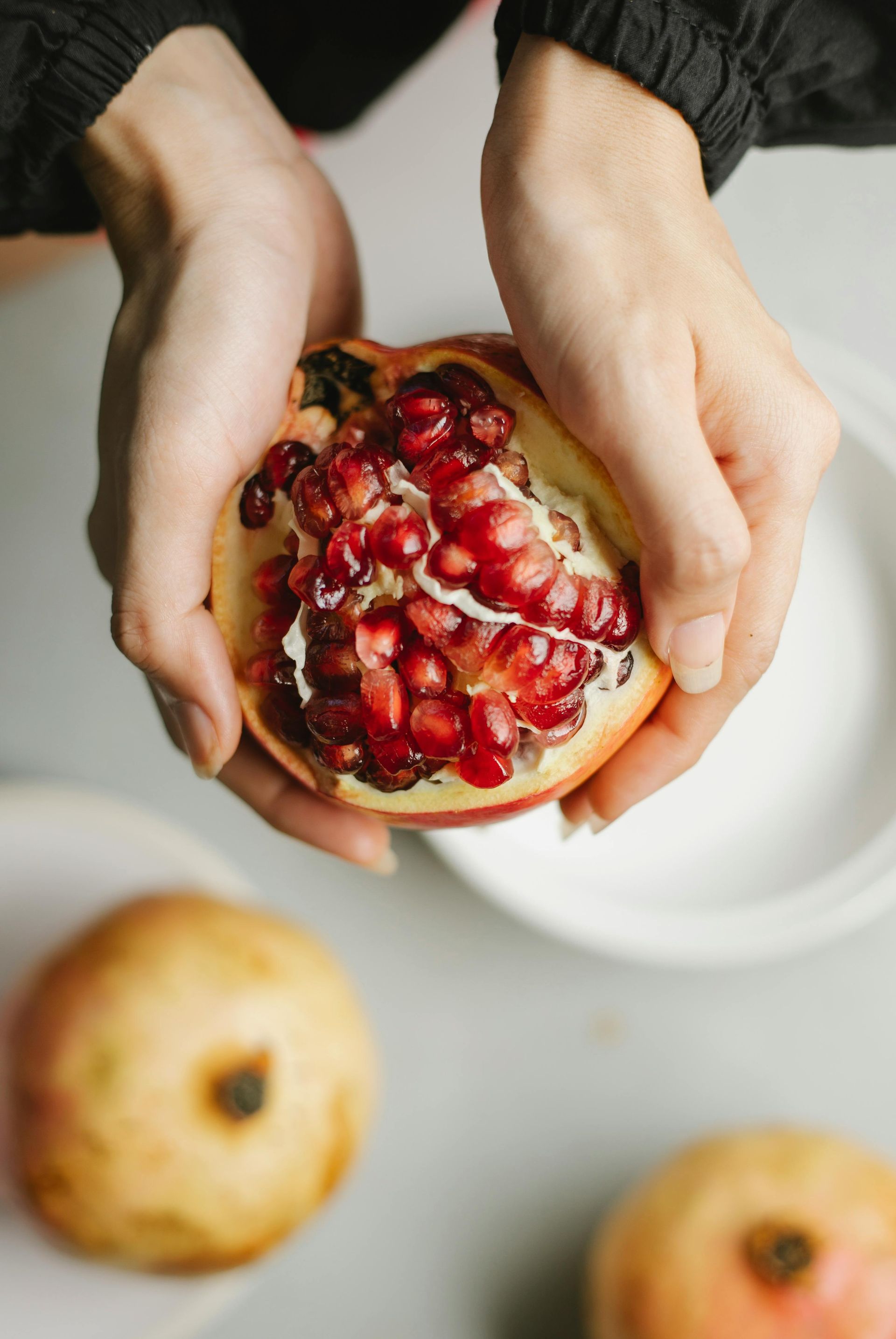 Hands holding a cut pomegranate, revealing red seeds. Two other pomegranates and white plates are visible on a light surface.