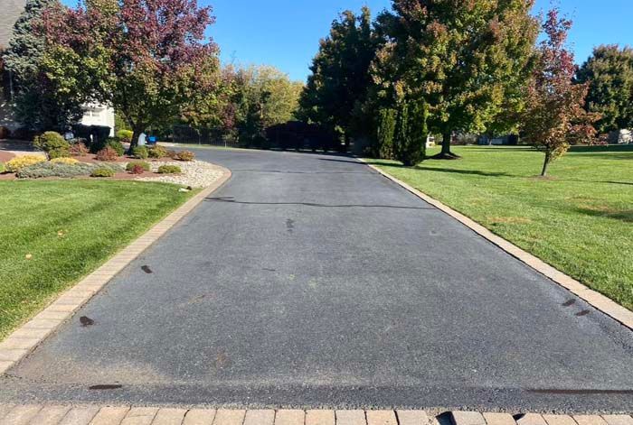 Asphalt driveway lined with brick, bordered by green lawn and trees under a blue sky.