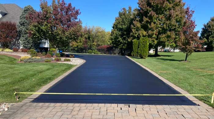 Newly paved black asphalt driveway with yellow caution tape, surrounded by green grass and trees.