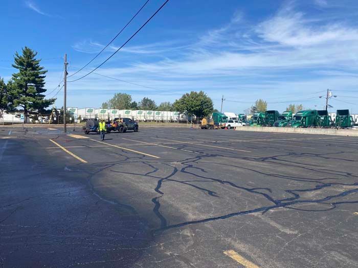 Asphalt parking lot with crack sealing underway; workers, equipment, semi-trucks, and utility poles visible.