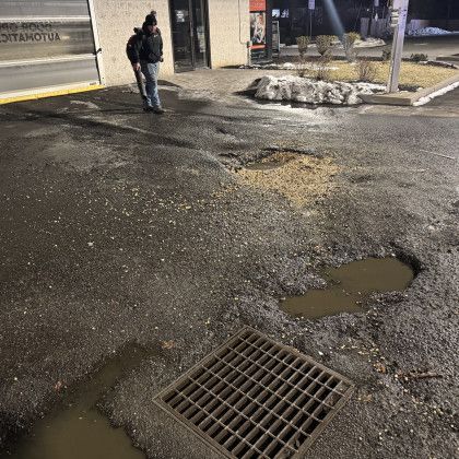 Man standing on pothole-ridden asphalt near a building entrance, drain, and small snow pile.